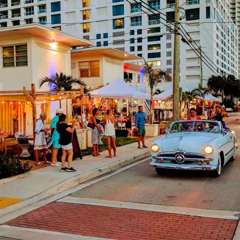 A white building with a tent and people outside, with a white car driving by.